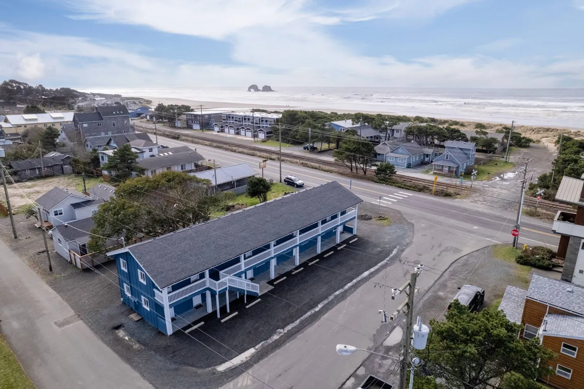 A tranquil exterior scene featuring the iconic blue building of the Inn with an expansive view of the ocean just steps away. The image includes beachgoers walking nearby and hints at surrounding local shops and homes, highlighting the lively beachfront atmosphere.