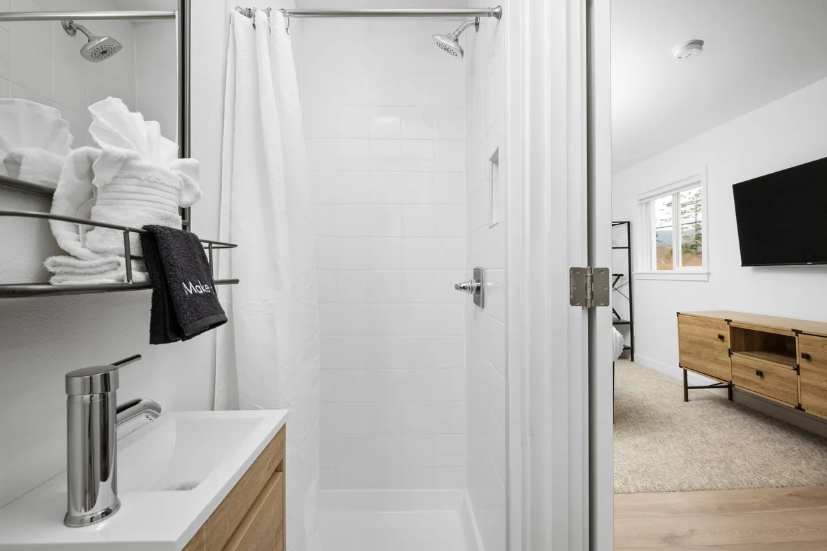 A serene, minimalistic bathroom with a spacious shower area, stacked fluffy towels on display, a sleek vanity sink, a large mirror, and a wall-mounted television. The scene is well-lit, emphasizing the clean lines and modern finishes that contribute to a spa-inspired atmosphere.