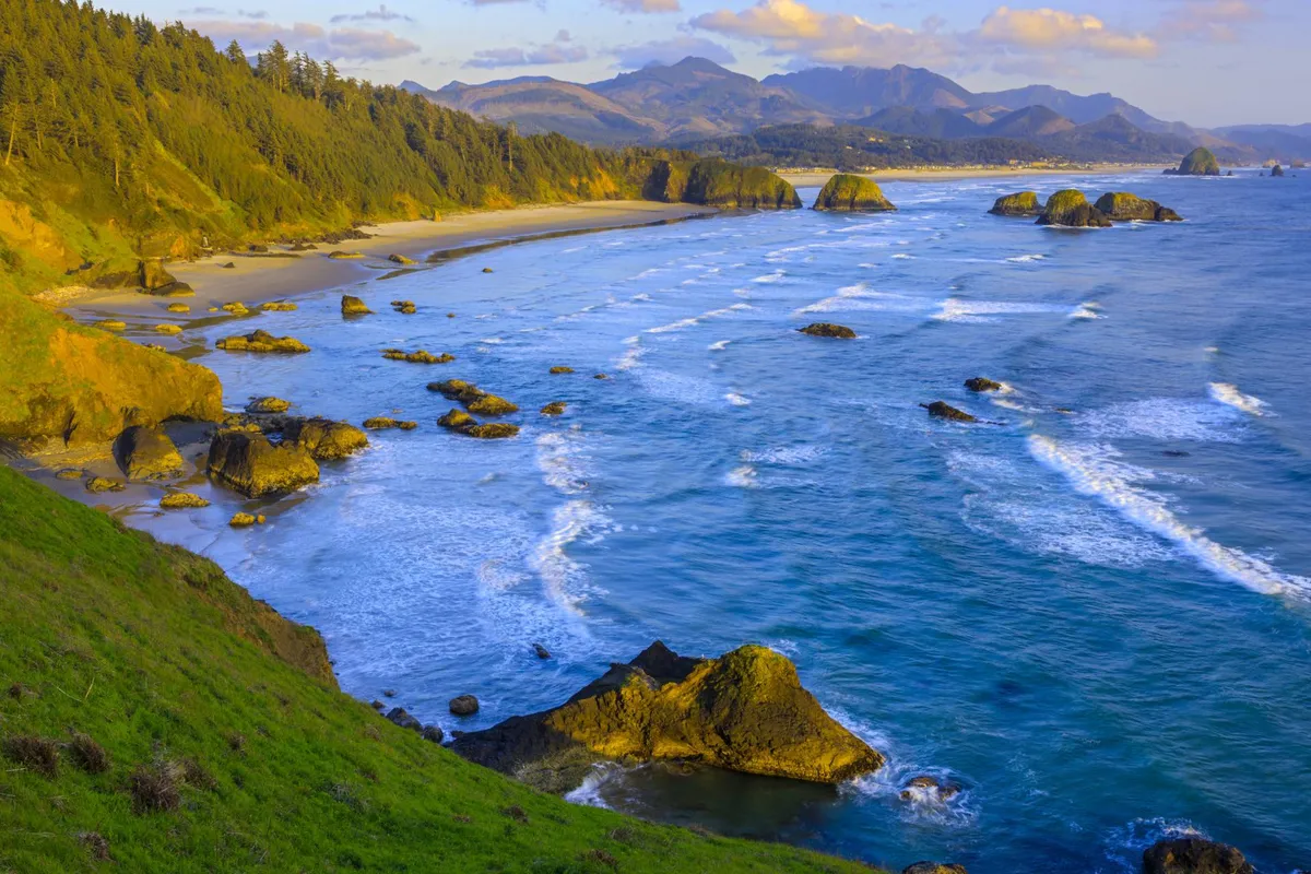 A scenic view of Rockaway Beach with gentle ocean waves rolling onto the sand, rugged rock formations, and distant mountains and forested hills providing a majestic backdrop. The peaceful scene captures the allure of sunset walks and time spent by the water.