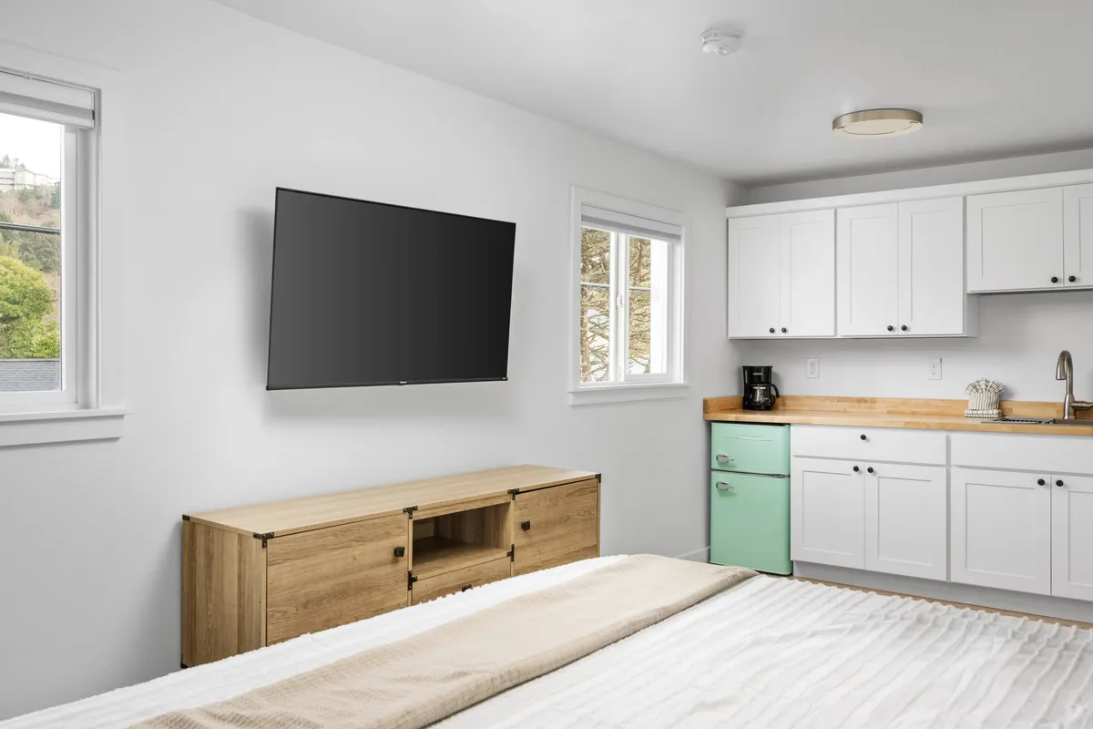 A broader view of the interior shows the living area with a comfortable bed, TV, and the kitchenette featuring the same green refrigerator. Natural light streams in, suggesting a gentle morning routine or relaxing evening in. The layout highlights how the living and kitchen spaces flow together, illustrating the room's adaptability for different moments of the day.